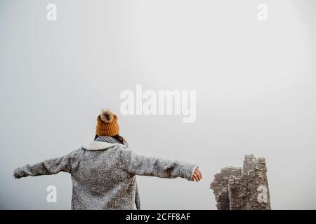 Rückansicht einer anonymen Frau in warmer Kleidung mit offenen Armen, die am bewölkten Tag im nebligen Park spazieren geht Stockfoto