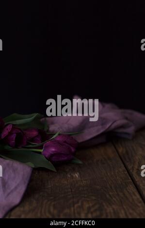 Bouquet von schönen dunkelvioletten Tulpen und Tuch auf gelegt Holz-Tischplatte vor schwarzem Hintergrund Stockfoto