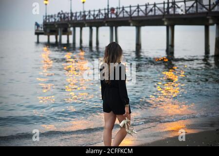 Junge Frau starrt auf das Meer mit Schuhen in der Hand in der Nähe pier drehte sich abends von der Kamera ab Stockfoto