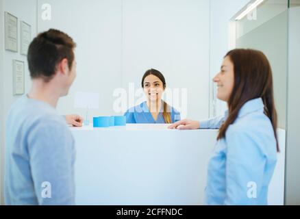 Seitenansicht eines lächelnden Gastes, der mit einem Mann an der Rezeption sprach Schreibtisch über begeisterte Administrator in der Zahnklinik Stockfoto