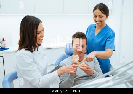 Begeisterter Patient im Stuhl mit Prothese und Zahnarzt mit Assistent In Uniform erklärenden Zahnstruktur Stockfoto