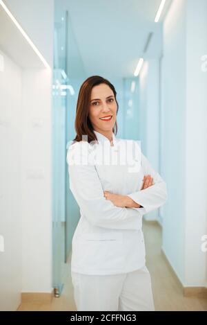 Fröhlich eleganter Arzt in weißer Uniform lächelt bei der Kamera mit Gekreuzte Arme im Lichtkorridor Stockfoto