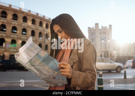 Junge ethnische Frau in stilvoller Kleidung, die die Karte beim Besuch der historischen Stadt an sonnigen Tagen untersucht Stockfoto