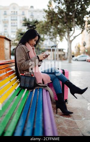 Seitenansicht Ganzkörper ethnische Dame mit Koffer sitzend auf rainbow Bank und Browsing Smartphone auf der Straße der Stadt Stockfoto