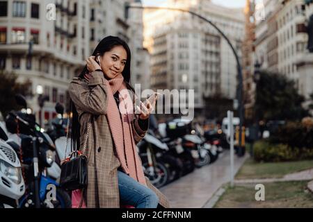 Glückliche asiatische Frau mit lächelndem Smartphone und Blick auf die Kamera, während sie auf dem Koffer auf dem Bürgersteig auf der Stadtstraße sitzt Stockfoto
