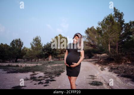 Glückliche Schwangere in legerem Kleid streichelte den Bauch, während sie am sonnigen Sommertag auf einem Pfad im Park mit grünen Bäumen und blauem Himmel stand Stockfoto