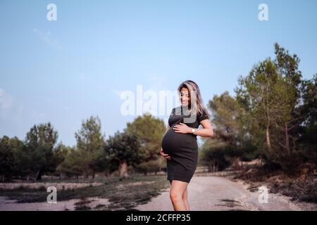 Glückliche Schwangere in legerem Kleid streichelte den Bauch, während sie am sonnigen Sommertag auf einem Pfad im Park mit grünen Bäumen und blauem Himmel stand Stockfoto