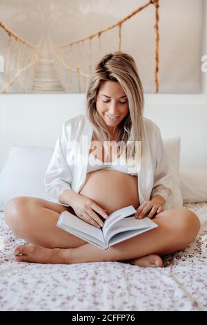 Inhalt blonde schwangere Frau trägt weiße elegante Kleidung zu Hause lesen Buch während sitzen mit gekreuzten Beinen auf dem Bett in hell Zimmer Stockfoto