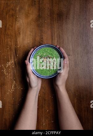 Draufsicht auf unkenntlich weibliche halten mit beiden Händen Keramik Kleine Schüssel frischen grünen Smoothie mit Zugabe von Kürbis Samen und Sesam gegen braunen Holztisch Stockfoto