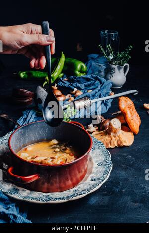 Beschnittene Hand einer nicht erkennbaren Person golding einen Löffel in der Nähe von A Schüssel mit köstlicher Suppe auf Tisch mit Rotkiefer gelegt Zum Abendessen werden Pilze und würzige grüne Chilischote serviert Stockfoto