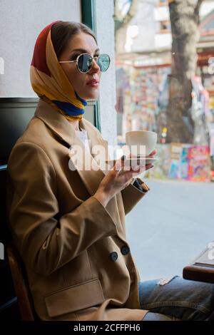 Seitenansicht der trendigen jungen Frau in Brillen und Schal Am Kopf Kaffee genießen, während Sie mit gekreuzten Beinen sitzen Cafeteria in der Nähe des Fensters am Nachmittag Stockfoto