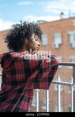 Nachdenkliche afroamerikanische Frau im karierten Hemd sitzend ruhenden Kopf Auf dem Balkon Metallzaun und wegschauen Stockfoto