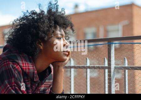 Nachdenkliche afroamerikanische Frau im karierten Hemd sitzend ruhenden Kopf Auf dem Balkon Metallzaun und wegschauen Stockfoto