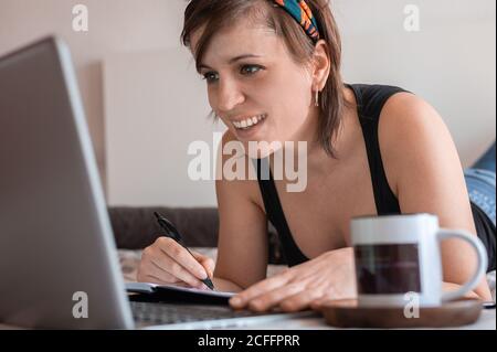 Seitenansicht der Crop Happy Woman in legerem Outfit, auf dem Bett mit Laptop und einer Tasse Kaffee liegend, während sie im Notizblock schrieb und sich am Wochenende erholte Stockfoto