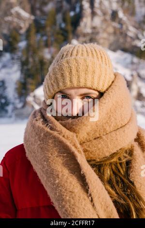 Junge aktive Frau in roter Jacke und gestricktem beigefarbenen Hut Eingewickelt in langen Wollschal neben verschneiten Bergen An sonnigen kalten Wintertag in Sibirien Russland Stockfoto