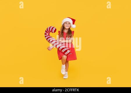 Ein niedliches kleines Mädchen Kind in einem ausgefallenen Kleid und einem Weihnachtsmann Hut hält eine aufblasbare Form eines Zuckerrohrs auf einem gelben Hintergrund. 2021 Neujahr con Stockfoto