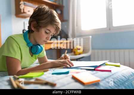 Nachdenklicher Schuljunge in legerer Kleidung und kabellosen Kopfhörern, die Musik genießen Und zeichnen mit Bleistiften während der Freizeit im Schlafzimmer Stockfoto