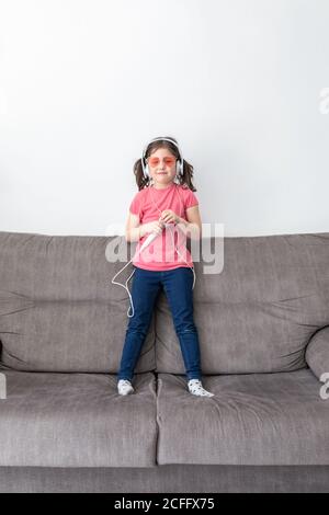 Fröhliches kleines Kind in rosa Brille und Kopfhörer Blick auf Kamera auf dem Sofa, während Spaß mit Mikrofon und vortäuscht Künstler im hellen Wohnzimmer der zeitgenössischen Wohnung Stockfoto