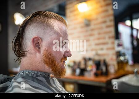 Seitenansicht des Rotschopf-Mannes, der in einem modernen Friseurladen sitzt Geschlossene Augen warten auf Friseur Stockfoto