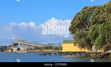 Panoramablick auf die Auckland Harbour Bridge, Auckland, Neuseeland, vom südlichen Ende gesehen. Rechts sind Bootshäuser und Pohutukawa-Bäume Stockfoto