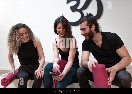 Glückliche junge Frau lächelt und zeigt den begeisterten Freunden mit Matten vor dem Yoga-Training im Studio Daten auf dem Smartphone Stockfoto