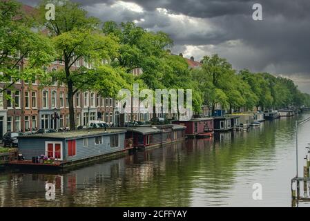 Ruhiges Wasser des Stadtkanals vor Sturm mit dunklen Wolken Über Gebäuden und grünen Bäumen in Amsterdam Stockfoto