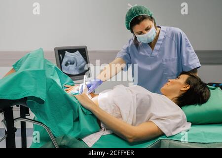 Ärztin in steriler Maske und blauem Handschuh mit Ultraschallscanner, während sie eine fröhliche Schwangere im Krankenhaus untersucht Stockfoto