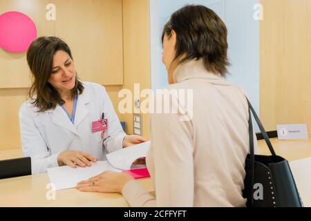 Lächelnder Spezialist in weißer Uniform und mit Namensschild Überprüfung Dokumente beim Gespräch mit einer nicht erkennbaren Patientin an der Rezeption In der Klinik Stockfoto