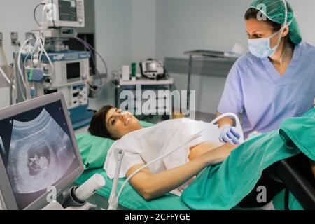Ärztin in steriler Maske und blauem Handschuh mit Ultraschallscanner, während sie eine fröhliche Schwangere untersucht und im Krankenhaus auf den Computerbildschirm schaut Stockfoto