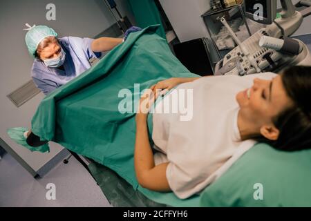 Ärztin in blauer Uniform und sterile Maske Untersuchung lächelnd Patient auf gynäkologischen Stuhl in der Fruchtbarkeitsklinik Stockfoto