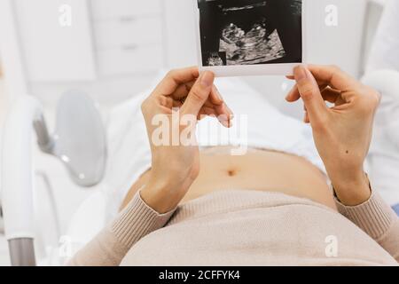 Beschnitten unkenntlich schwangere Frau inspiziert Sonogramm Bild während liegend Bett auf der Station der modernen Fruchtbarkeitsklinik Stockfoto