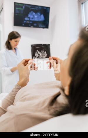 Nicht erkennbare schwangere Frau inspiziert Sonogramm Bild, während sie auf dem Bett liegen Auf der Station der modernen Fruchtbarkeit Klinik Stockfoto