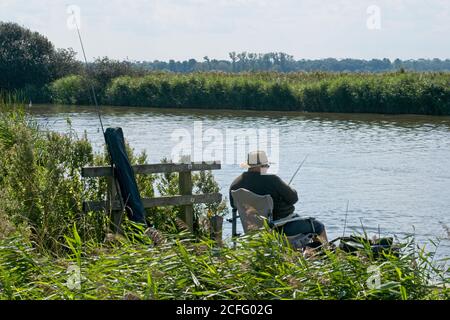 Grobfischerei auf den Norfolk Broads der Mann sitzt entspannt am Schilfufer und angelt mit Rute. Schilf und Felder gegenüber. Leichter, wolkig. Ansicht schließen. Stockfoto