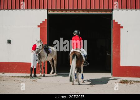 Rückansicht einer anonymen jungen Teenager-Frau in Jockey-Helm und Jacke, die auf einem Pferd neben dem Stalleingang steht Stockfoto