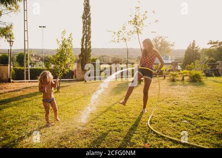 Ganzkörper fröhlich Teenager Hosing kleine Mädchen mit sauberem Wasser Beim Spaß auf Rasen im Hof zusammen Stockfoto