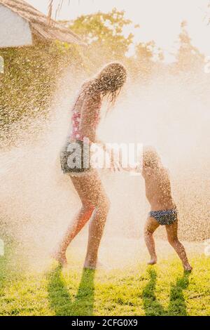 Ganzkörper anonyme Teenager und kleines Mädchen laufen und spielen In Tropfen des Spritzens sauberes Wasser, während Spaß in Garten zusammen Stockfoto