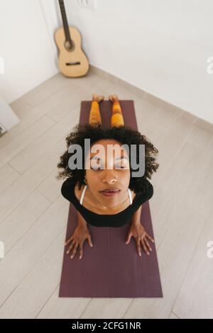 Von oben Gesicht der afroamerikanischen attraktiven jungen Frau sitzt in Yoga-Pose mit geschlossenen Augen auf Matte in hellen Raum Stockfoto