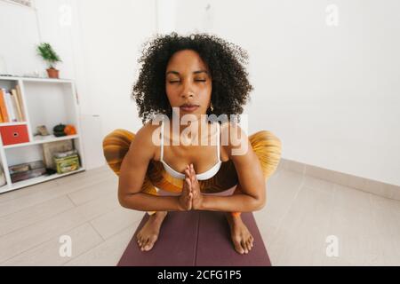 Afroamerikanisch attraktive junge Frau, die Yoga-Pose durchführt, mit geschlossenen Augen, die sich zu Hause auf einer Matte hocken Stockfoto