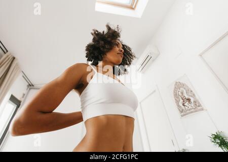 Von unten afroamerikanische attraktive junge Frau, die Yoga-Haltung mit gefalteten Händen hinter dem Rücken in hellen Raum Stockfoto