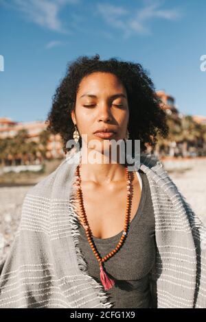 Afroamerikanische attraktive junge Frau, die mit grauem Karos bedeckt ist und an hellen Tagen in einer Lotusyogatage am Sandstrand meditiert Stockfoto