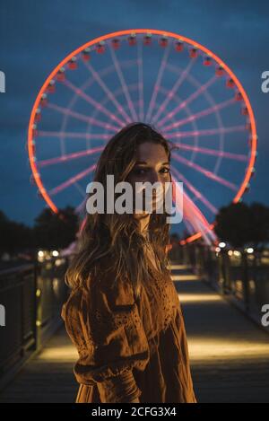 Junge Touristenfrau, die auf dem beleuchteten Pier steht und die Kamera mit dem leuchtenden Riesenrad im Hintergrund in Montreal anschaut Stockfoto