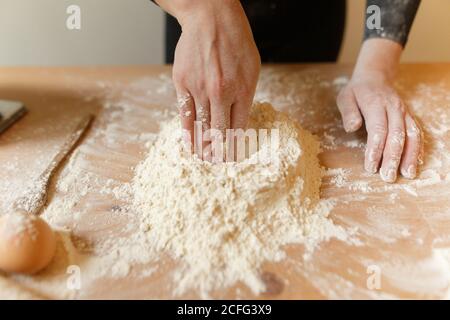 Anonyme Person in schwarzen Kleidern, die eine Vertiefung in Mehlmasse machen Mit der Hand zum Hinzufügen von Eiern beim Kochen von Teig für Pasta Zu Hause Stockfoto