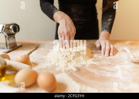 Anonyme Person in schwarzen Kleidern, die eine Vertiefung in Mehlmasse machen Mit der Hand zum Hinzufügen von Eiern beim Kochen von Teig für Pasta Zu Hause Stockfoto