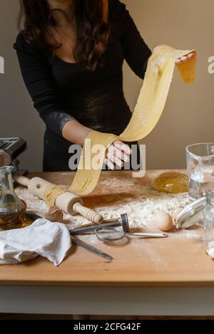 Anonyme Dame in schwarzem Kleid hält fein gerollten Nudelteig In den Händen und stehend neben unordentlichen Tisch beim Kochen Italienisch Gericht zu Hause Stockfoto