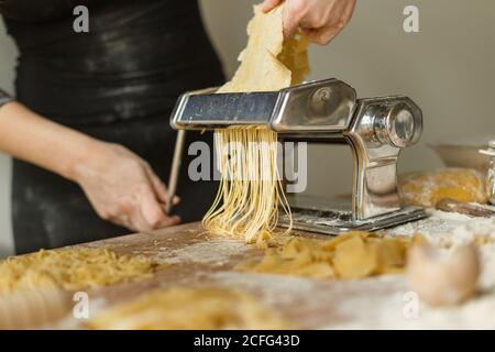 Crop anonyme Frau in schwarzem Kleid schneiden Teig in dünne Streifen mit Pasta-Maschine während der Zubereitung verschiedener Formen von Pasta zu Hause Stockfoto