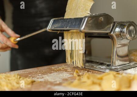 Crop anonyme Frau in schwarzem Kleid schneiden Teig in dünne Streifen mit Pasta-Maschine während der Zubereitung verschiedener Formen von Pasta zu Hause Stockfoto