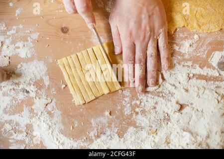 Faceless Kochen Schneid Teig in dünne Streifen mit Messer auf Holztisch mit Mehl bestreut, während die Pasta tagsüber zubereitet wird Stockfoto