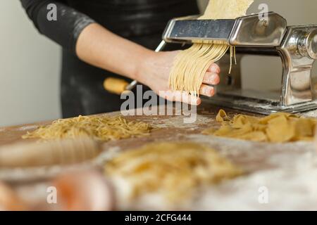 Crop anonyme Frau in schwarzem Kleid schneiden Teig in dünne Streifen mit Pasta-Maschine während der Zubereitung verschiedener Formen von Pasta zu Hause Stockfoto