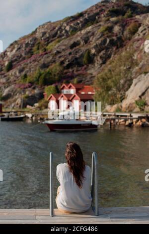 Rückansicht von anonymen weiblichen Touristen in lässiger warmer Kleidung Sitzen auf Holzboardwalk und bewundern Sie die ruhige Aussicht auf das Boot Und Häuser Stockfoto