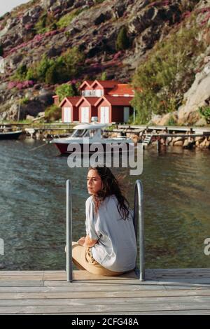 Rückansicht der weiblichen Touristen in legerer Kleidung sitzend auf Hölzerne Promenade mit Blick auf die ruhige Aussicht auf das Boot und Häuser Stockfoto